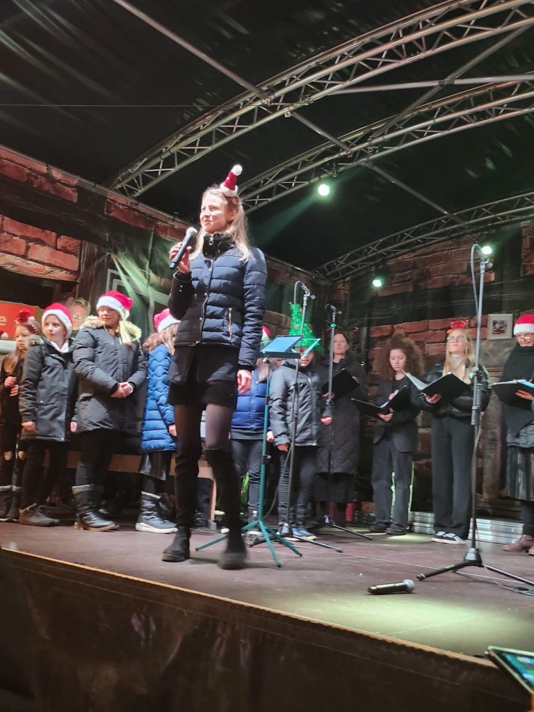 Magdalena Ehlers die Chorleiterin von Chor Cantiamo steht dem Publikum zugewandt auf der Bühne des Weihnachtsmarktes in Wolfsburg und spricht in ein Mikrofon. Hinter ihr stehen Kinder und junge Erwachsene, die Chormappen in den Händen tragen und Weihnachtsschmuck auf dem Kopf. Die Bühne ist geziert mit einem Tannenbaum und bunten Lichtern, die weihnachtliche Stimmung vermitteln