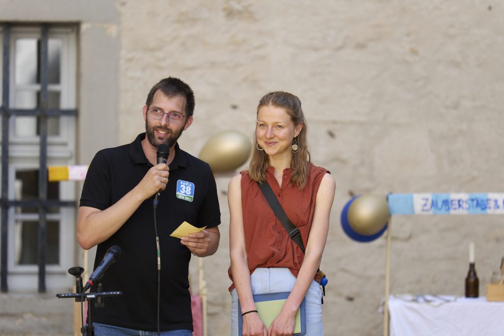 Chorleiterin Magdalena Ehlers wird von einem Radiomoderator von Radio38 interviewt. Sie stehen kurz vor der Aufführung des Musiktheaters der Chorakademie ft. Theater & Tanz Wolfsburg.
Beide lächeln. Im Hintergrund sind Luftballons und Marktstände zu sehen.
Es ist der Jugendchor der Chorakademie Wolfsburg. Sie spielen ein Theaterstück, das sie selbst geschrieben haben. Leistung Magdalena Ehlers. Foto: Tim Dalmann. Ort: Schloss Wolfsburg. Chor: Chor Cantiamo e.V.