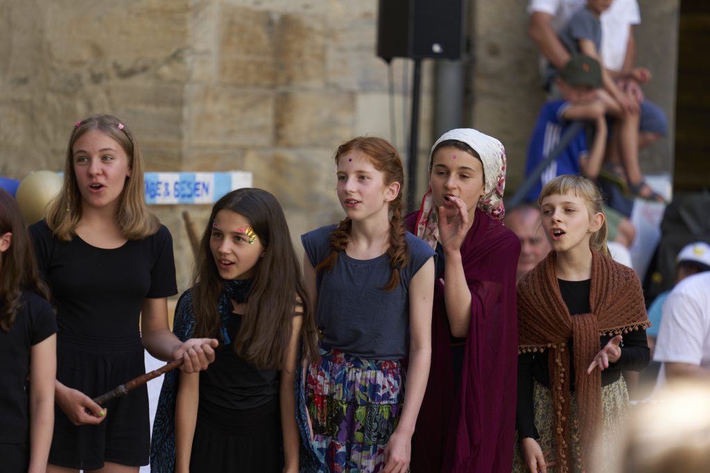 Fünf (5) Mädchen stehen hintereinander und schauen alle interessiert in eine Richtung. Sie singen. Es ist der Jugendchor der Chorakademie Wolfsburg. Sie spielen ein Theaterstück, das sie selbst geschrieben haben. Leistung Magdalena Ehlers. Foto: Tim Dalmann. Ort: Schloss Wolfsburg. Chor: Chor Cantiamo e.V.