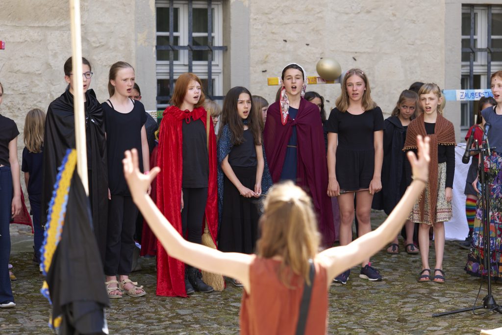 Ein Chor singt auf einer Theaterbühne. Man sieht die dirigirerenden Arme der Chorleiterin Magdalena Ehlers.
Es ist der Jugendchor der Chorakademie Wolfsburg. Sie spielen ein Theaterstück, das sie selbst geschrieben haben. Leistung Magdalena Ehlers. Foto: Tim Dalmann. Ort: Schloss Wolfsburg. Chor: Chor Cantiamo e.V.