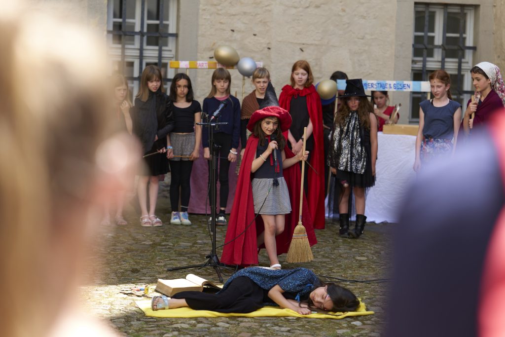 Ein Mädchen in Umhang und Hexenbesen hält ein Mikrofon in der Hand. sie hat einen Zauberhut auf dem Kopf. Im Hintergrund steht der Kinderchor der Chorakademie Wolfsburg. Vom Chor Cantiamo e.V.  Im Hintergrund sind Marktstände zu sehen, an denen Luftballons hängen. Sie spielen ein Theaterstück mit der Chorakademie Wolfsburg fest Theater & Tanz im Schloss Wolfsburg