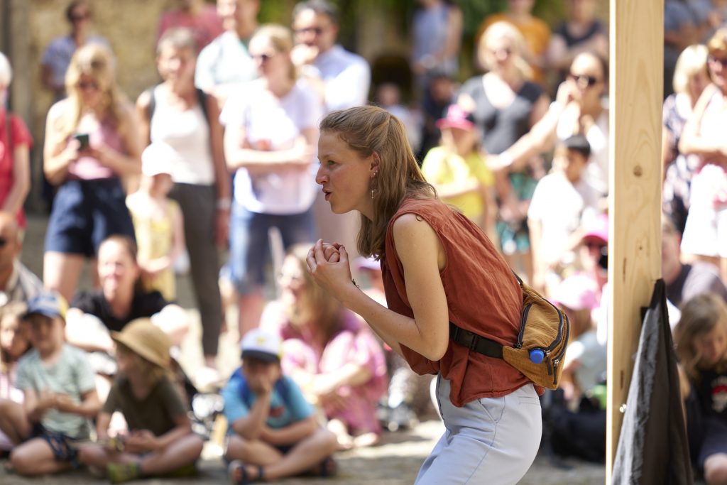 Chorleiterin Magdalena Ehlers schaut intensiv und dirigiert den Chor. Im Hintergrund ist ein zahlreiches Publikum in der Sonne zu sehen. Es ist der Jugendchor der Chorakademie Wolfsburg. Sie spielen ein Theaterstück, das sie selbst geschrieben haben. Leistung Magdalena Ehlers. Foto: Tim Dalmann. Ort: Schloss Wolfsburg. Chor: Chor Cantiamo e.V.