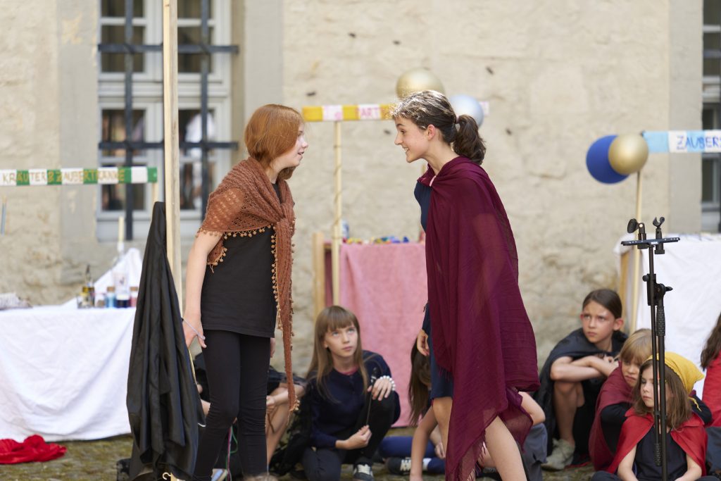 zwei Mädchen in Umhängen stehen sich gegenüber und schauen sich überrascht an. Im Hintergrund sind Marktstände zu sehen, an denen Luftballons hängen. Sie spielen ein Theaterstück mit der Chorakademie Wolfsburg fest Theater & Tanz im Schloss Wolfsburg